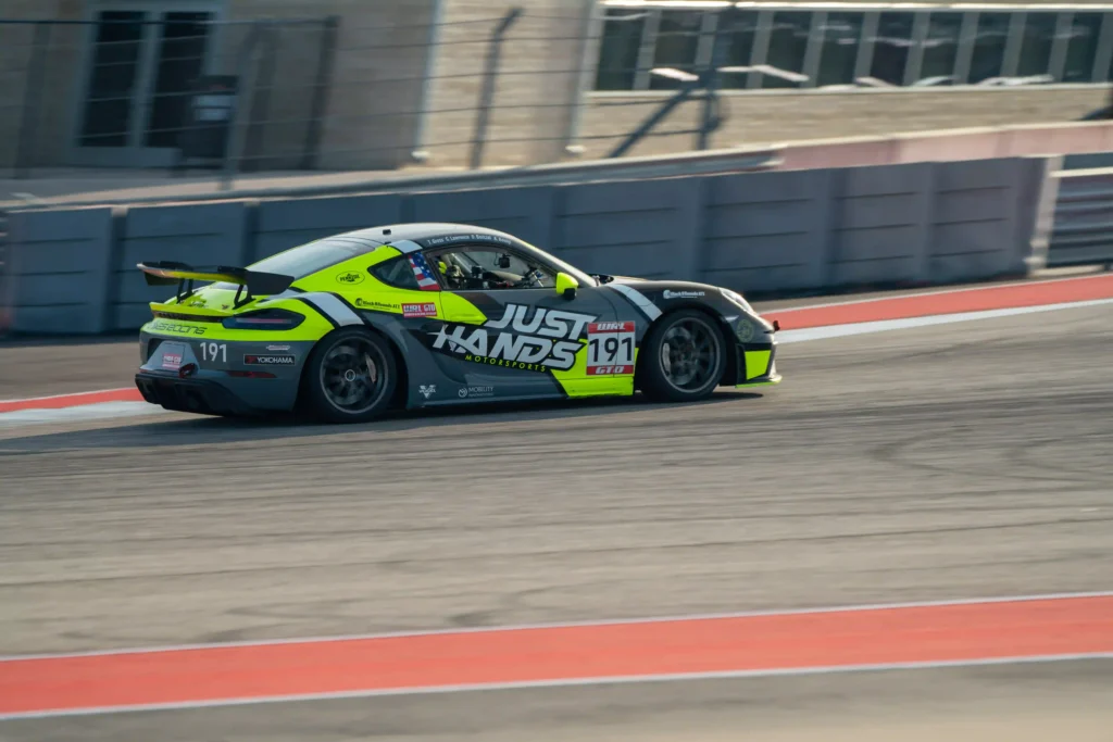 A race car is speeding down a track, featuring a sleek design and bright color scheme. The car's body is primarily dark grey with neon yellow and white accents. Large text on the side reads "Just Hands Motorsports" with the number "191" prominently displayed. The car has various sponsor logos, including Yokohama tires, and is equipped with a rear wing spoiler for aerodynamic performance. The background shows a concrete barrier and blurred structures, emphasizing the car's motion as it navigates a corner on the track.