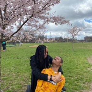 A young couple (sister and disabled brother) is enjoying a moment under a blooming cherry blossom tree in a park. The woman, with long braided hair, is standing behind the man, who is seated in a wheelchair and wearing a bright yellow shirt. She lovingly wraps her arms around him while they gaze into each other's eyes, smiling softly. The setting is a green open space, possibly a sports field, with overcast skies and a few more cherry trees in the background. The mood is warm and caring, with nature's beauty surrounding them.