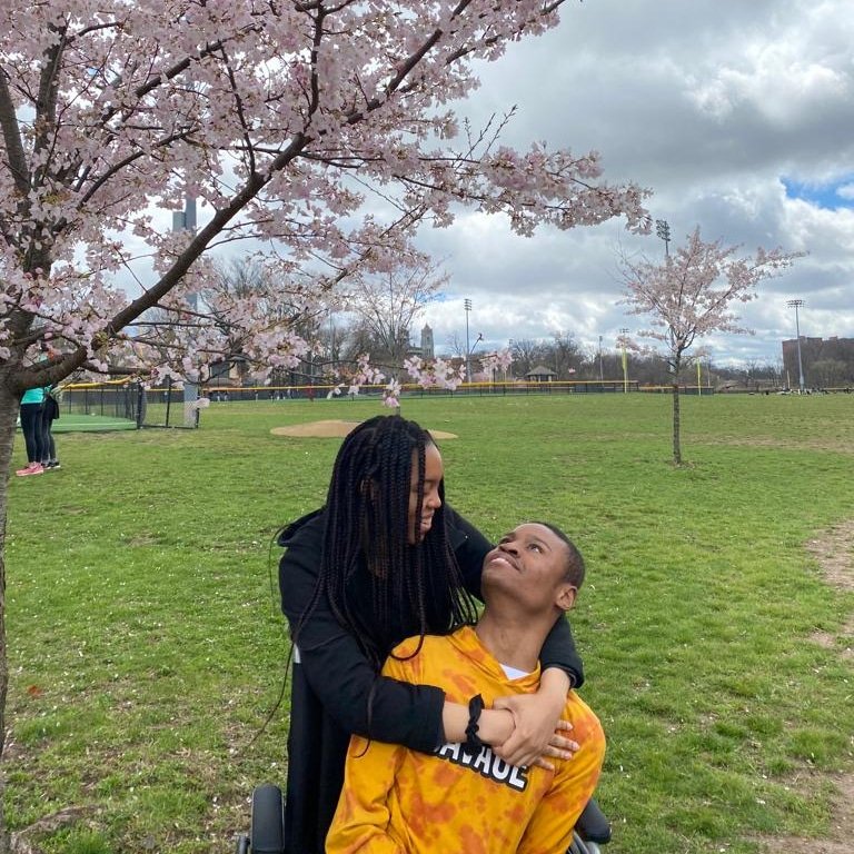 A young couple (sister and disabled brother) is enjoying a moment under a blooming cherry blossom tree in a park. The woman, with long braided hair, is standing behind the man, who is seated in a wheelchair and wearing a bright yellow shirt. She lovingly wraps her arms around him while they gaze into each other's eyes, smiling softly. The setting is a green open space, possibly a sports field, with overcast skies and a few more cherry trees in the background. The mood is warm and caring, with nature's beauty surrounding them.