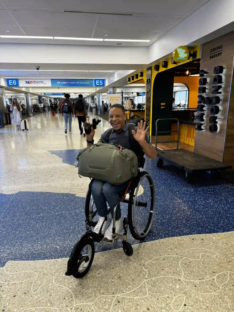 A man in a wheelchair is smiling and waving at the camera while navigating through an airport terminal. He is wearing glasses and a grey shirt, with a large backpack on his lap. The wheelchair has an additional front wheel attachment, likely for easier mobility. The terminal around him shows gate signs (E5 and E6), and there are other travelers in the background. A sign for "Harvest Grounds" coffee and bakery is visible on the right. The atmosphere appears lively and friendly, with bright lighting and a clean, modern design.