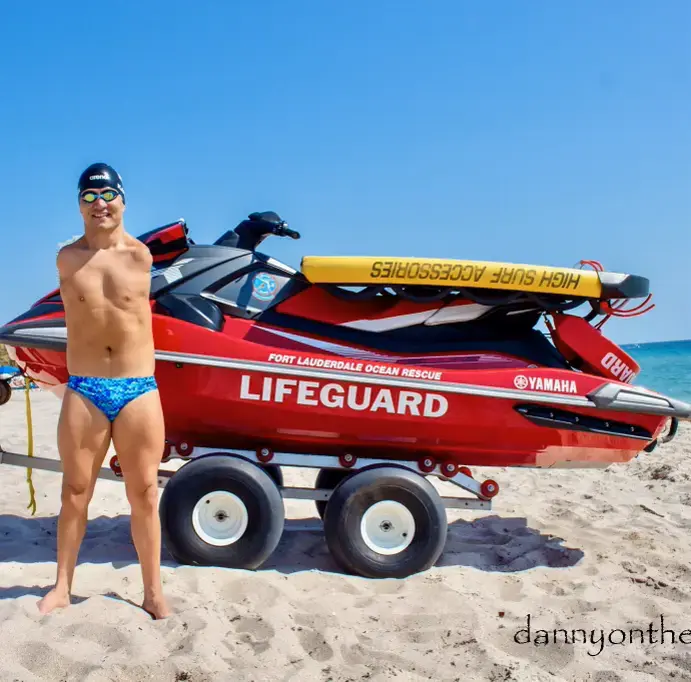 The image shows a lifeguard standing on a sandy beach next to a red rescue vehicle, specifically a jet ski mounted on a wheeled trailer. The lifeguard is wearing blue swim briefs with a patterned design, swim goggles on their head, and appears to be smiling. The rescue vehicle has the text "FORT LAUDERDALE OCEAN RESCUE" and "LIFEGUARD" prominently displayed on the side, along with a "YAMAHA" logo. The ocean is visible in the background under a clear blue sky.
