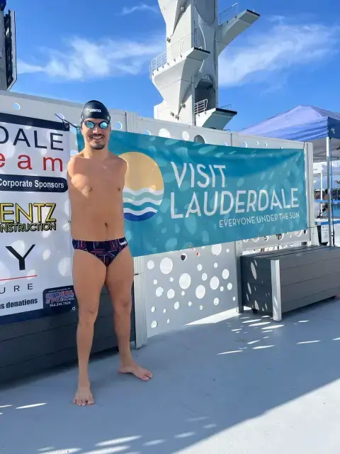 The image shows a male swimmer with no arms standing in front of a promotional banner that reads "Visit Lauderdale - Everyone Under the Sun." The swimmer is wearing a black swim cap with reflective goggles and a black and red patterned swim brief. He is standing barefoot by the side of a pool, with a diving platform visible in the background. The setting appears to be a sunny day at an outdoor aquatic venue, possibly during a competition or event. The swimmer is smiling confidently at the camera.