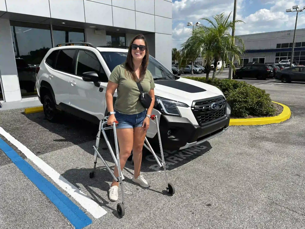 A woman with long brown hair, wearing sunglasses, a green t-shirt, blue shorts, and white sneakers, is standing outside in front of a white SUV. She is smiling and using a walker for mobility. The car is parked in a handicap-accessible parking spot, marked by blue lines. The scene takes place in a sunny area, with palm trees and a building in the background, possibly a car dealership.