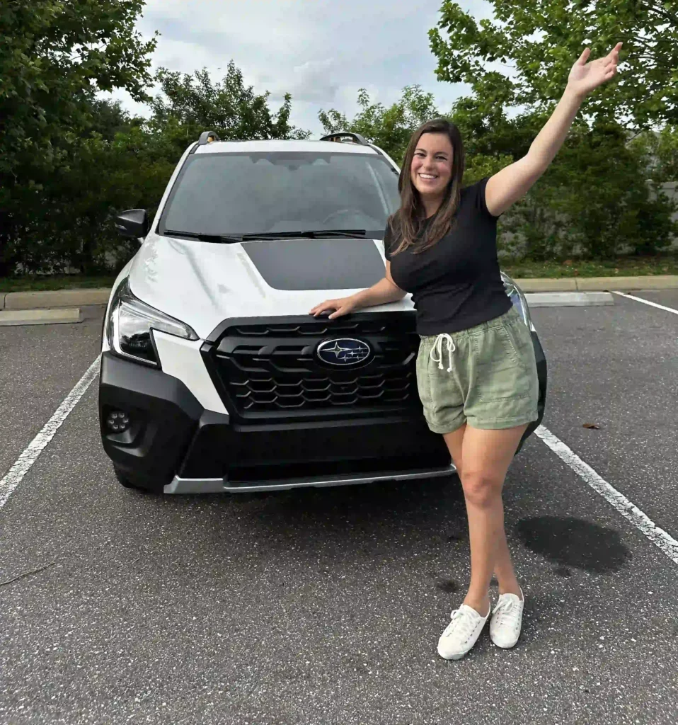 A woman is standing in front of a Subaru vehicle, smiling and posing with her right arm extended upwards and her left hand resting on the car. She is wearing a black t-shirt, green shorts with a drawstring, and white sneakers. The car is parked in an outdoor lot, with trees and greenery visible in the background. The Subaru has a black grille with a silver logo prominently displayed. The sky is slightly cloudy, giving the scene a calm and pleasant vibe.