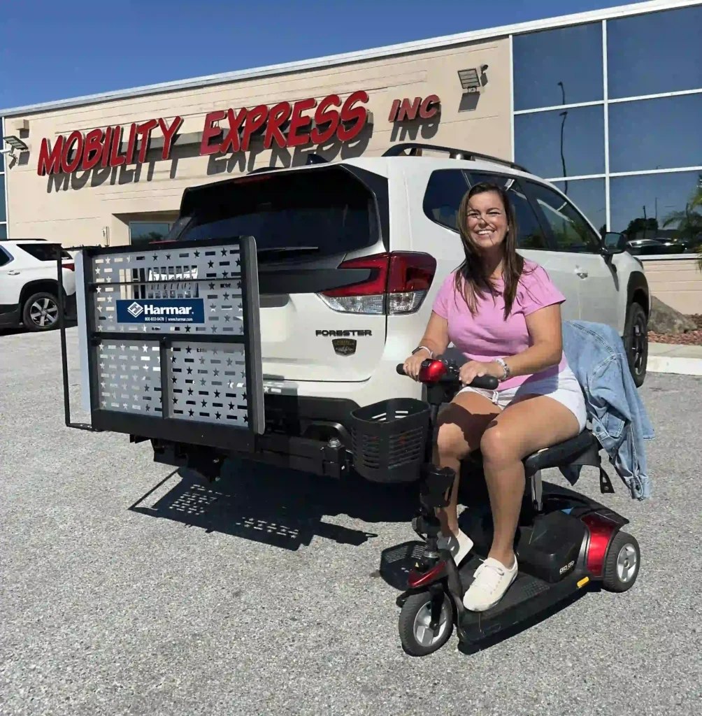 A woman is sitting on a mobility scooter in front of a white SUV, which has a "Harmar" mobility lift attached to the rear. She is wearing a light pink shirt, white shorts, and white sneakers, smiling at the camera with a casual and cheerful expression. There is a denim jacket draped over the back of her seat. The SUV is parked in front of a store with a sign that reads "Mobility Express Inc." The weather appears to be sunny and clear, as evidenced by the bright lighting and clear blue sky.