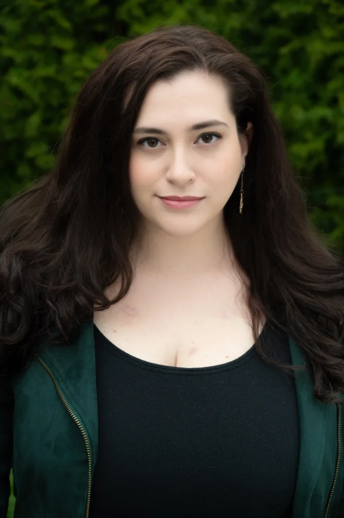 A young woman with long, dark brown hair, wearing a dark green jacket over a black top, stands against a blurred, green foliage background. She has a calm expression with neutral makeup, gazing directly at the camera.