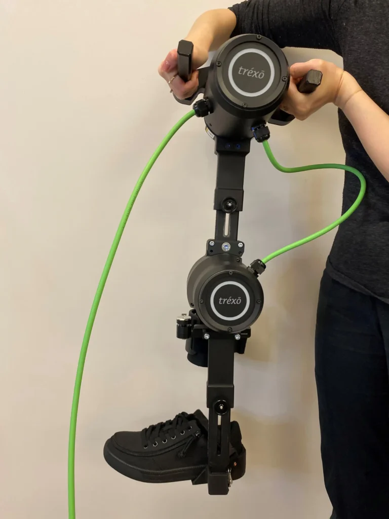 The image shows a person holding a robotic leg device, branded with the name "Trexo." The device appears to be an exoskeleton or robotic-assisted mobility system, featuring mechanical joints and a foot holder fitted with a black sneaker. Green cables are connected to the system, possibly for power or control purposes. The person is gripping handles at the top of the device, giving the impression that they are either adjusting or preparing the device for use.