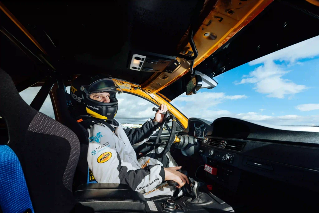The image shows a race car driver seated inside a car, wearing a helmet and racing suit. The driver is holding the steering wheel with one hand and appears to be focused on the road ahead. The car's interior is stripped down for racing purposes, with exposed metal and wiring, as well as a roll cage for safety. The dashboard is minimalist, with a visible gear shifter and various switches. The driver seems ready to drive on a racetrack, with a clear blue sky and some clouds visible through the windshield.