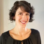 A woman is smiling in this portrait-style photo. She has short, curly dark hair and is wearing a black blouse with lace detailing on the shoulders. She is also wearing silver dangling earrings. The background is plain, emphasizing her friendly and approachable expression. The lighting is soft, giving the image a warm and welcoming feel.