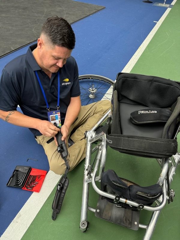 A man in a US Open shirt kneels beside a wheelchair, carefully working on its parts with tools spread nearby. He appears focused as he adjusts or repairs a component of the wheelchair. A set of hex keys lies on the floor next to him, indicating a detailed maintenance task.