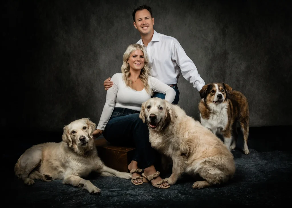 The image shows a couple posing with their three large dogs. The woman, seated in the front, is smiling and wearing a white top and jeans, with one arm resting on a Golden Retriever beside her. Her husband stands behind her with his hand gently placed on her shoulder, wearing a white shirt and jeans. Two other dogs, one Golden Retriever and one brown-and-white mixed breed, are positioned on either side of the couple, creating a warm family portrait. The setting has a neutral, dark backdrop, emphasizing the family's bond and connection with their pets.