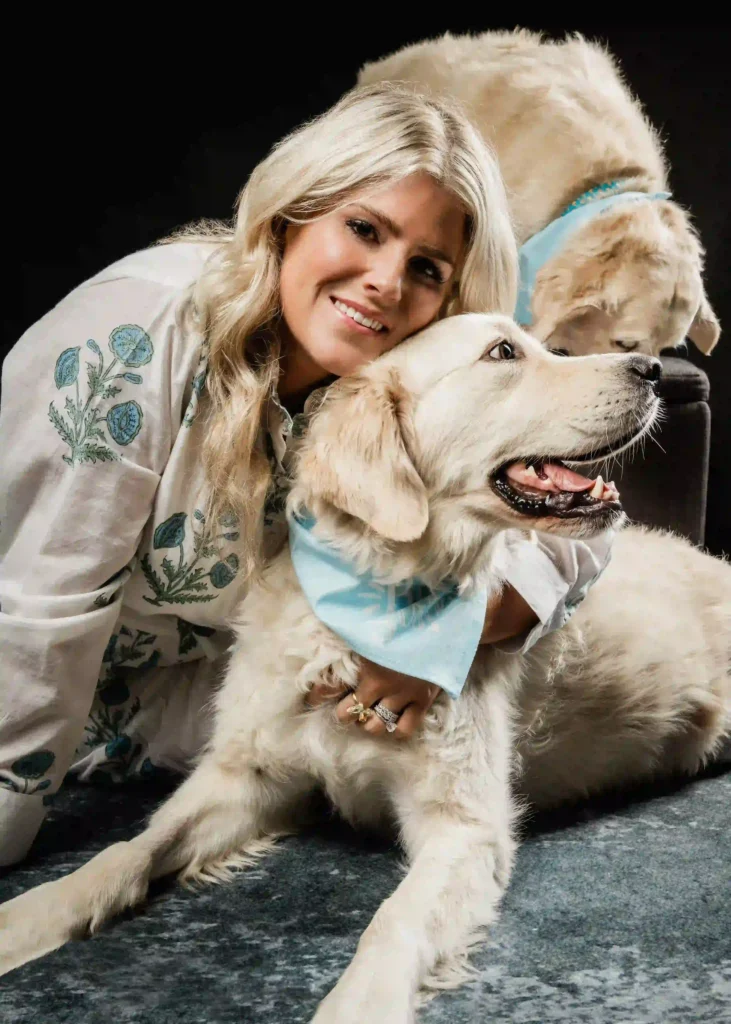 A woman with long blonde hair is smiling warmly as she hugs a Golden Retriever wearing a light blue bandana. She is dressed in a white top with blue floral embroidery. Another Golden Retriever is in the background, partially out of focus, resting its head on a chair. The setting appears to be cozy and intimate, with both dogs looking relaxed and happy. The scene radiates affection and a deep bond between the woman and her pets, highlighting a moment of joy and connection.