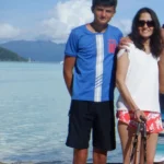 A photo of a young boy and a woman standing on a beach. The boy is wearing a blue t-shirt with the number 10 and the word "France" on it, along with black shorts. He has short dark hair and is standing with his arms behind his back. The woman beside him is wearing a white top and red floral shorts, with sunglasses on. She is holding two walking canes, and the beach stretches behind them with calm waters and distant mountains under a partly cloudy sky.