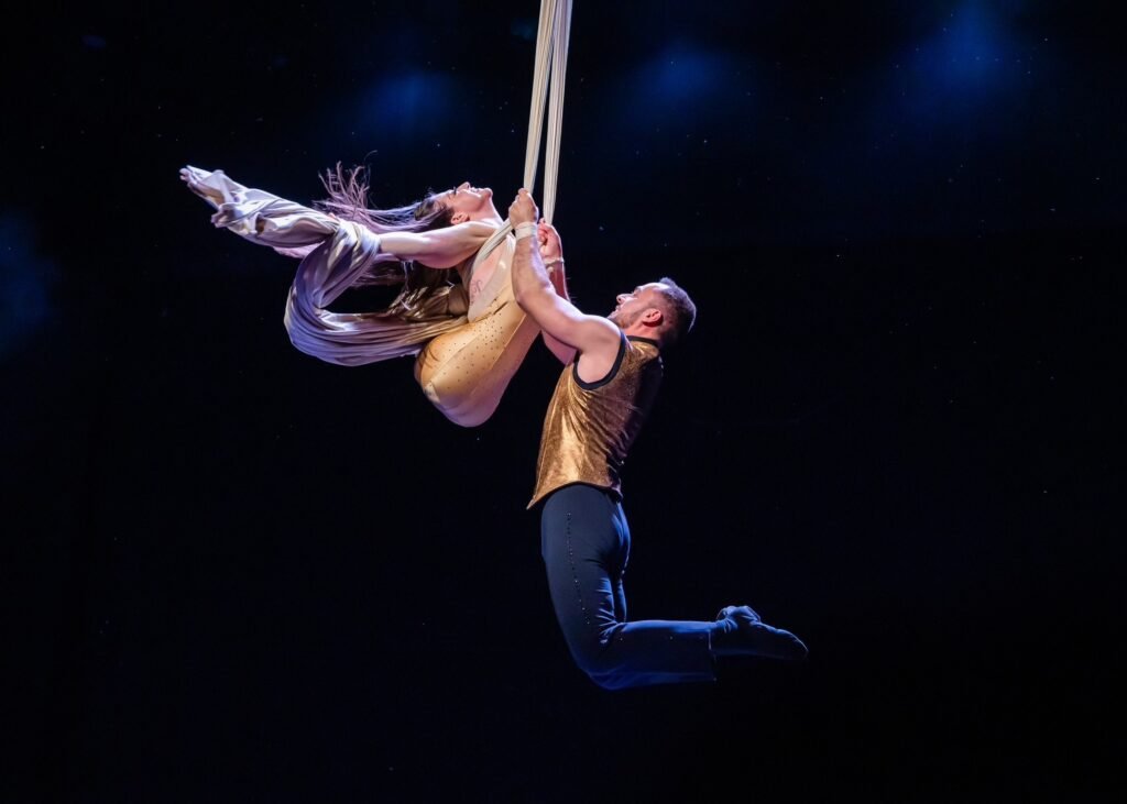 Two aerial performers gracefully suspended in mid-air on white silk ribbons, executing a synchronized acrobatic routine. The female performer, with no legs, dressed in a flowing outfit, is held securely by the male performer, who is wearing a gold vest and dark pants. The background is dark with spotlights illuminating the scene, highlighting the strength and balance required for their performance.