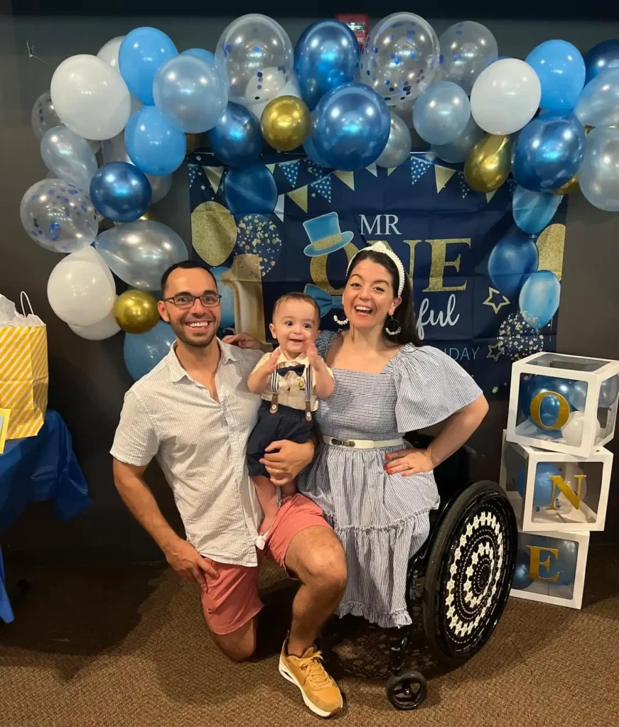 Photo of a joyful family celebrating a child's first birthday. The father is kneeling on one knee, smiling broadly, while holding their young son in the center. The mother, who uses a wheelchair, is also smiling and wearing a blue and white striped dress. She has her arm around her son, who is dressed in a stylish outfit with suspenders. Behind them is a balloon arch in blue, white, and gold, and a banner reading 'Mr. ONEderful.' Stacked blocks to the side spell out 'ONE,' adding to the festive atmosphere.