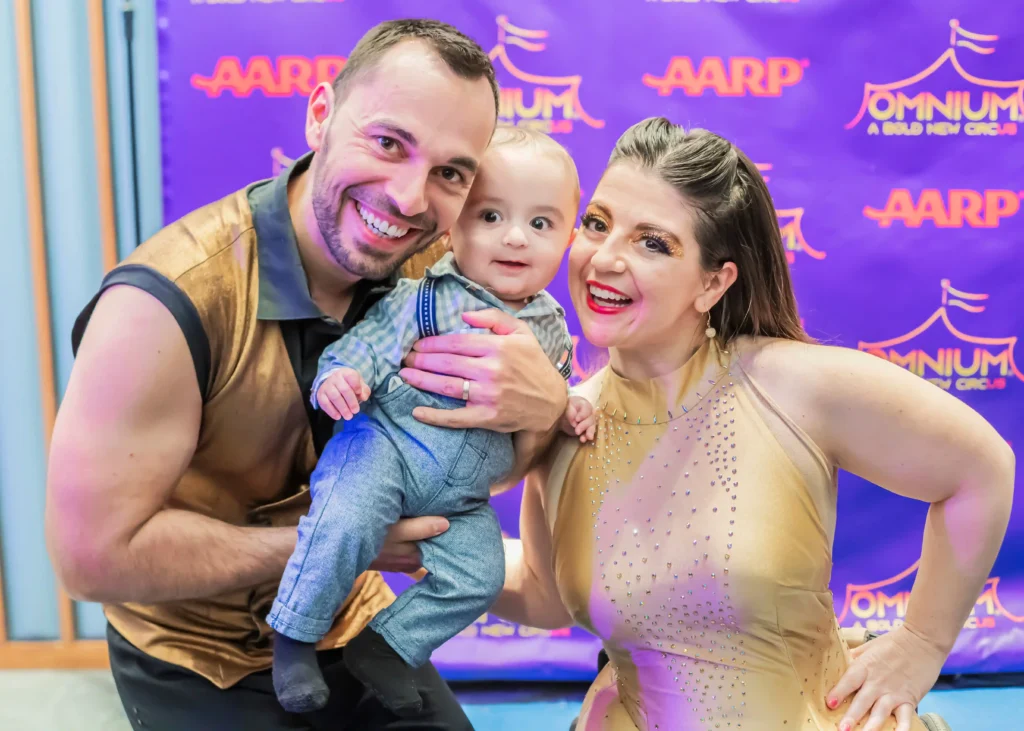 Smiling man and woman holding a baby at a vibrant event. The man, wearing a sleeveless gold and black top, beams at the camera while holding the baby, who is dressed in a plaid shirt and overalls. The woman, wearing a sparkling gold outfit, smiles brightly as she stands beside them. A purple backdrop featuring 'AARP' and 'Omnium: A Bold New Circus' logos is visible in the background