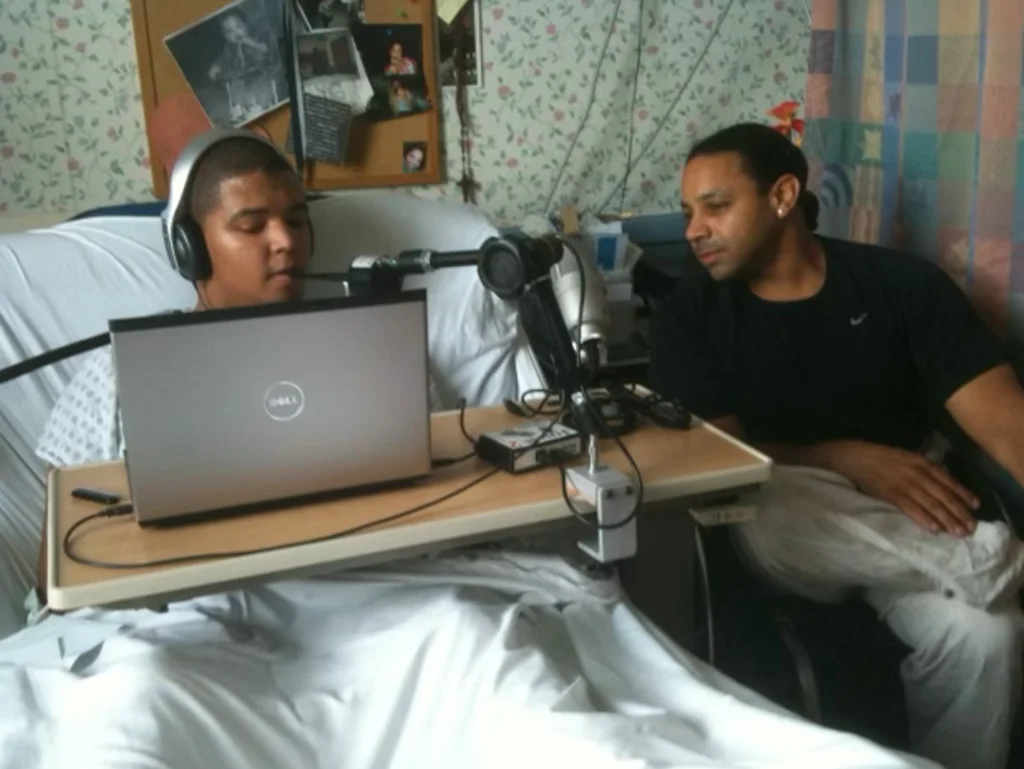 The image shows two men in what appears to be a hospital setting. The man on the left is lying in a hospital bed, using a laptop placed on a rolling table in front of him. He is wearing headphones and speaking into a microphone attached to the table, suggesting he might be recording something, possibly a podcast or an audio message. There is some audio equipment on the table as well. The man on the right, seated next to the bed, appears to be watching or listening attentively. The environment features hospital room decor, with floral-patterned wallpaper and a curtain, likely for privacy.