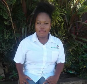 A young woman with natural hair pulled up in a puff, wearing a white collared shirt with an 'Academy' logo on the left side. She is seated outdoors against a background of lush green foliage, looking at the camera with a neutral expression.