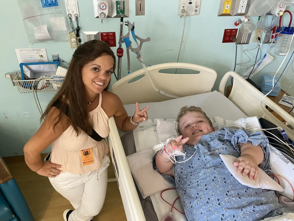The image shows Kristen DeAndrade standing next to a hospital bed, smiling and flashing a peace sign with her fingers. Beside her in the bed is a young child, also smiling and giving a peace sign, while wearing a hospital gown and connected to medical equipment. The setting appears to be a hospital room, with various medical devices visible in the background. The moment captured radiates warmth and positivity, emphasizing Kristen’s supportive presence and her connection with the child.