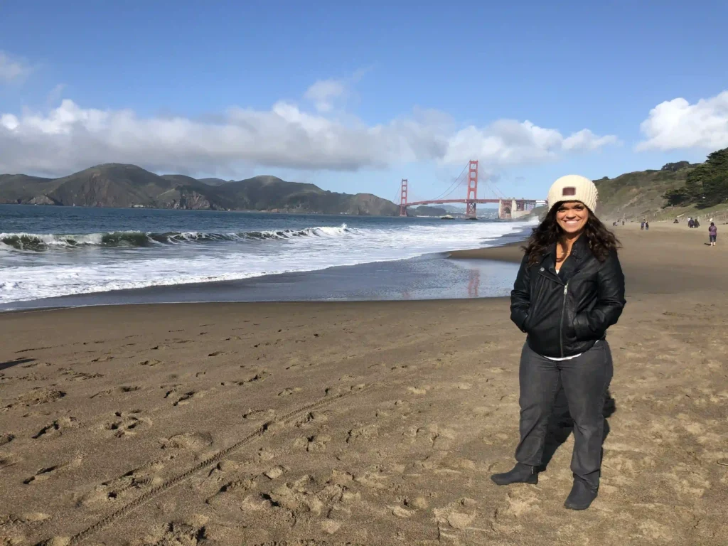 A woman is standing on a sandy beach with the Golden Gate Bridge visible in the background. The day is sunny with clear skies and a few clouds, and waves gently crash against the shore. The woman is smiling, wearing a black leather jacket, gray pants, black boots, and a cream-colored knit beanie. She has her hands in her jacket pockets, and there are footprints visible in the sand around her. The landscape features the iconic bridge, hills in the distance, and a few people strolling along the beach.