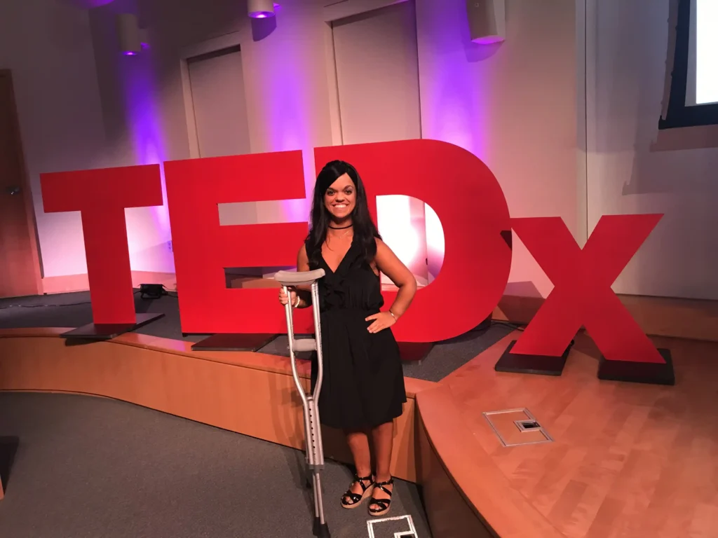 Kristen DeAndrade is standing in front of large, red TEDx letters on a stage. She is smiling confidently and holding a crutch, dressed in a black sleeveless dress with sandals. The lighting is warm, with purple tones illuminating the background, and Kristen appears ready to deliver a talk or presentation. This image captures her poised and inspiring presence at a TEDx event, highlighting her advocacy and storytelling.