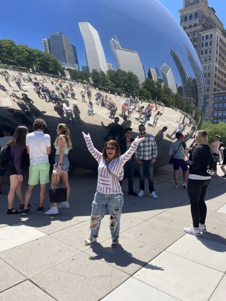 A woman stands in the foreground of a vibrant outdoor scene, posing excitedly with arms raised in front of "The Bean" (Cloud Gate) sculpture in Chicago's Millennium Park. Her striped top, ripped jeans, and sandals create a casual, cheerful vibe. The reflective surface of the sculpture mirrors the crowd and iconic Chicago skyline, including several tall buildings and green trees under a clear blue sky. Several other visitors are also gathered around, some chatting and taking photos, creating a lively, social atmosphere.