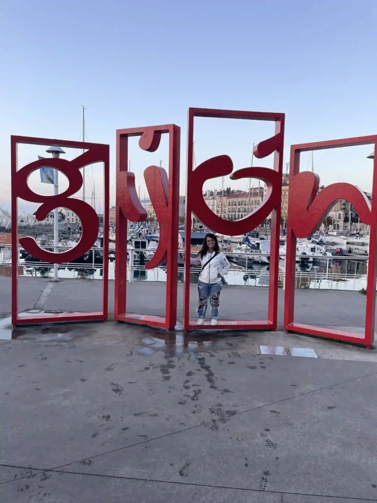 The image shows a person standing behind large red metal frames shaped into the letters of the word "Gijón." The letters are stylized and curved, resembling a modern or artistic font. The background reveals a marina with boats and masts, suggesting the location is near a harbor. The person in the photo is positioned in front of the middle letter, with a mix of casual attire, including jeans and a jacket. The sky appears clear, with soft lighting indicating early morning or late afternoon. The scene has a relaxed, coastal vibe.