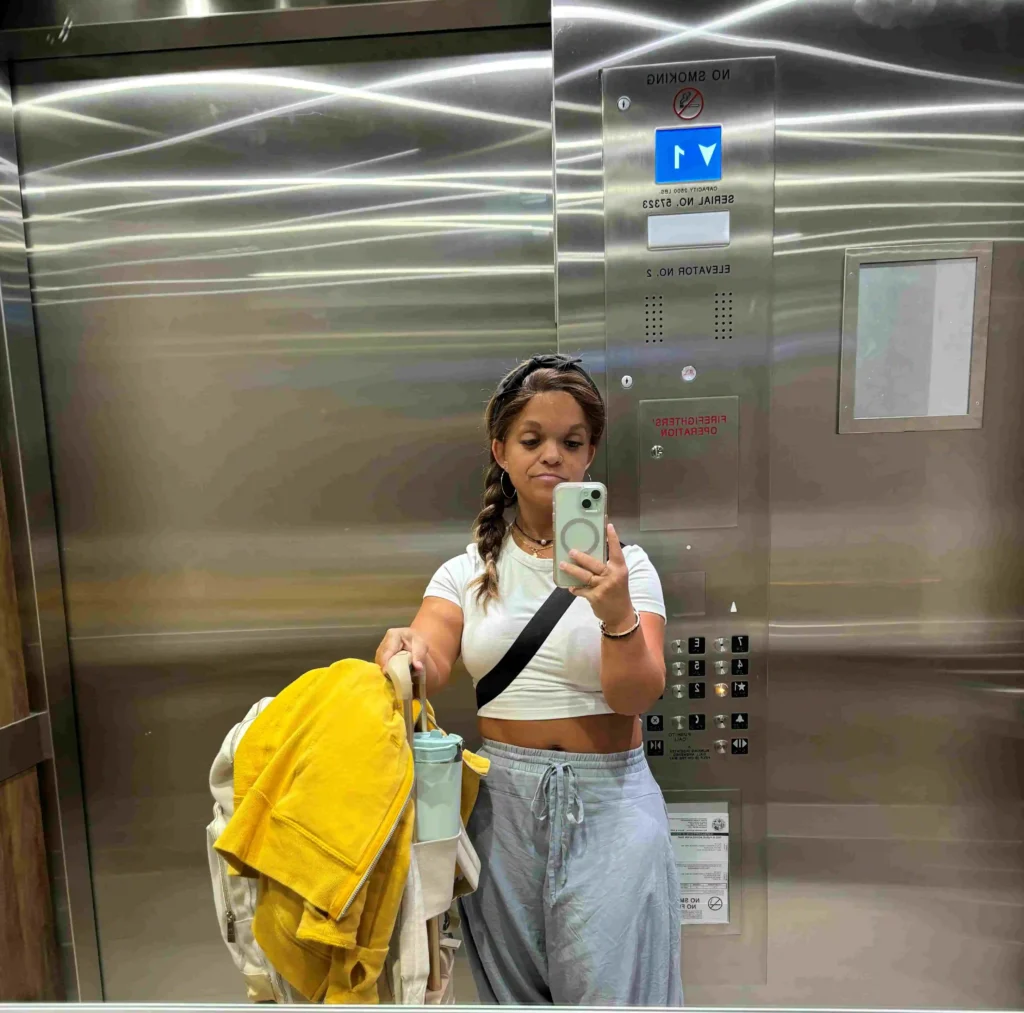 A woman is standing in an elevator, taking a mirror selfie. She is wearing a white cropped T-shirt, light blue pants, and a black crossbody bag. Her hair is braided and she is holding a large beige backpack with a yellow jacket and a light blue water bottle attached. The elevator has stainless steel walls with a no-smoking sign, buttons, and indicators visible. The lighting in the elevator creates reflections and patterns on the walls.