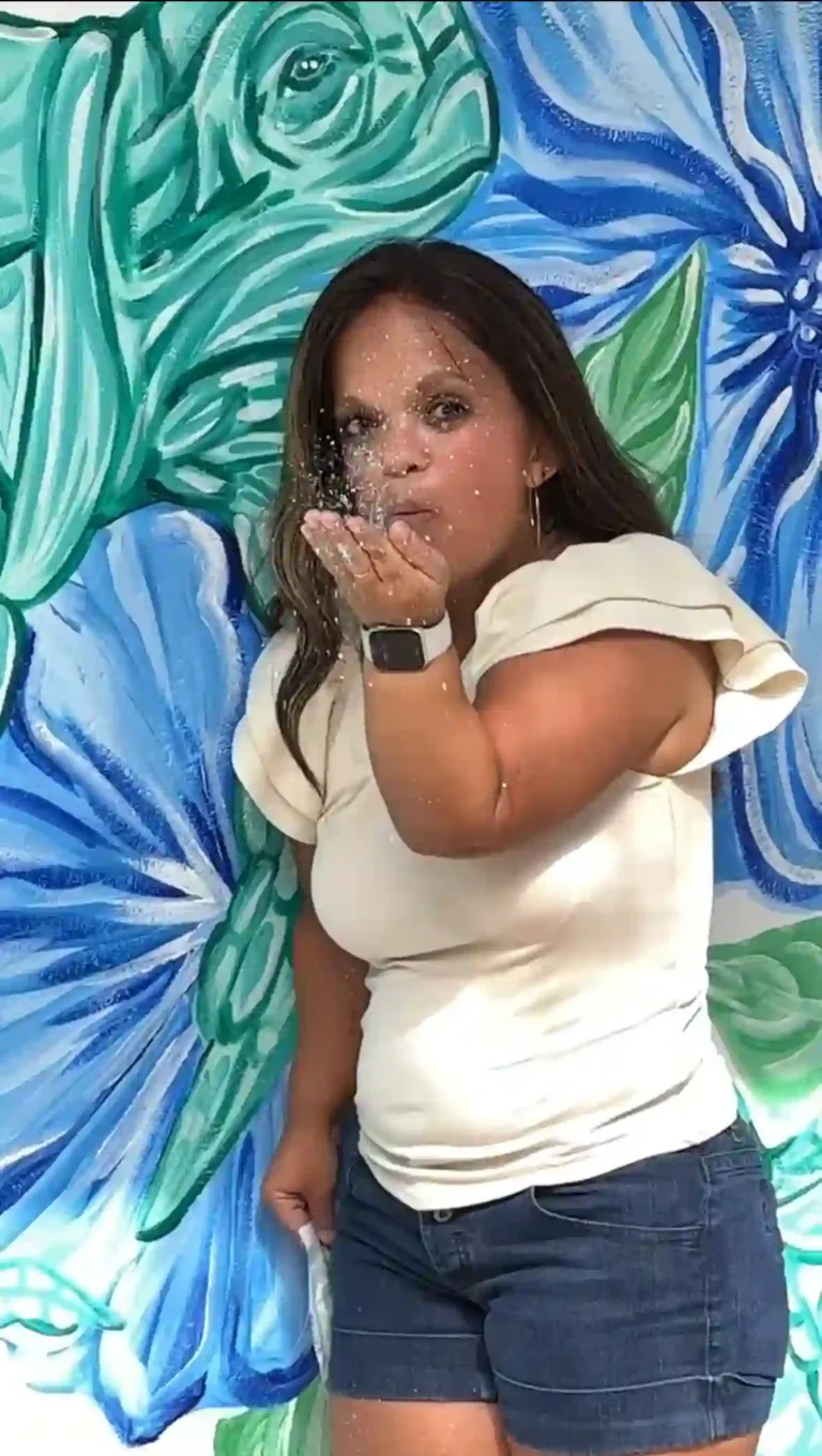 The image shows a woman standing in front of a vibrant mural that features large, abstract floral patterns in shades of blue and green. She is blowing glitter from her hand toward the camera, creating a dynamic, sparkly effect. The woman has long brown hair and is wearing a cream-colored top with short ruffled sleeves and dark denim shorts. She also has a smartwatch on her wrist. The background and the glitter give the image a lively, artistic vibe.