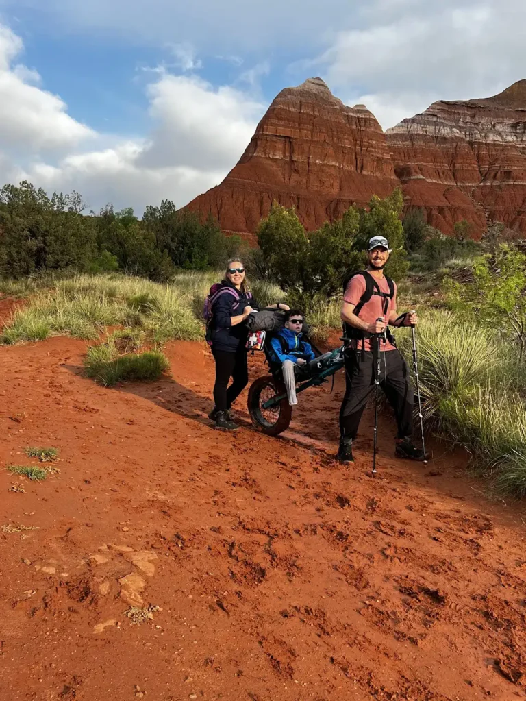 A family hiking in a desert-like environment is shown in the image. The landscape features reddish rock formations, with one prominent, layered mesa or butte in the background. The ground is a vibrant red dirt, and the surrounding area has some patches of green vegetation. In the foreground, two adults, a man and a woman, are seen pushing a special type of off-road wheelchair or cart with a young person seated inside. The man is wearing hiking gear, including sunglasses, a cap, trekking poles, and a backpack, while the woman, also in hiking gear, is wearing sunglasses, a backpack, and holding the cart. The child in the wheelchair is dressed in outdoor clothing and sunglasses, comfortably seated as they enjoy the scenery. The scene conveys a sense of adventure, inclusion, and family bonding during outdoor activities.