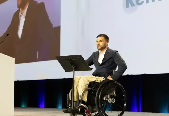 The image shows a person using a wheelchair (Leo Samarelli), positioned on a stage in front of a podium. The individual is delivering a speech or presentation and is dressed in a professional outfit consisting of a dark blazer and light-colored pants. The background features a large screen that displays a partial view of the same person projected behind them. The setting appears to be a formal or public speaking event, possibly a conference or keynote address. The person is confidently engaged with the audience.