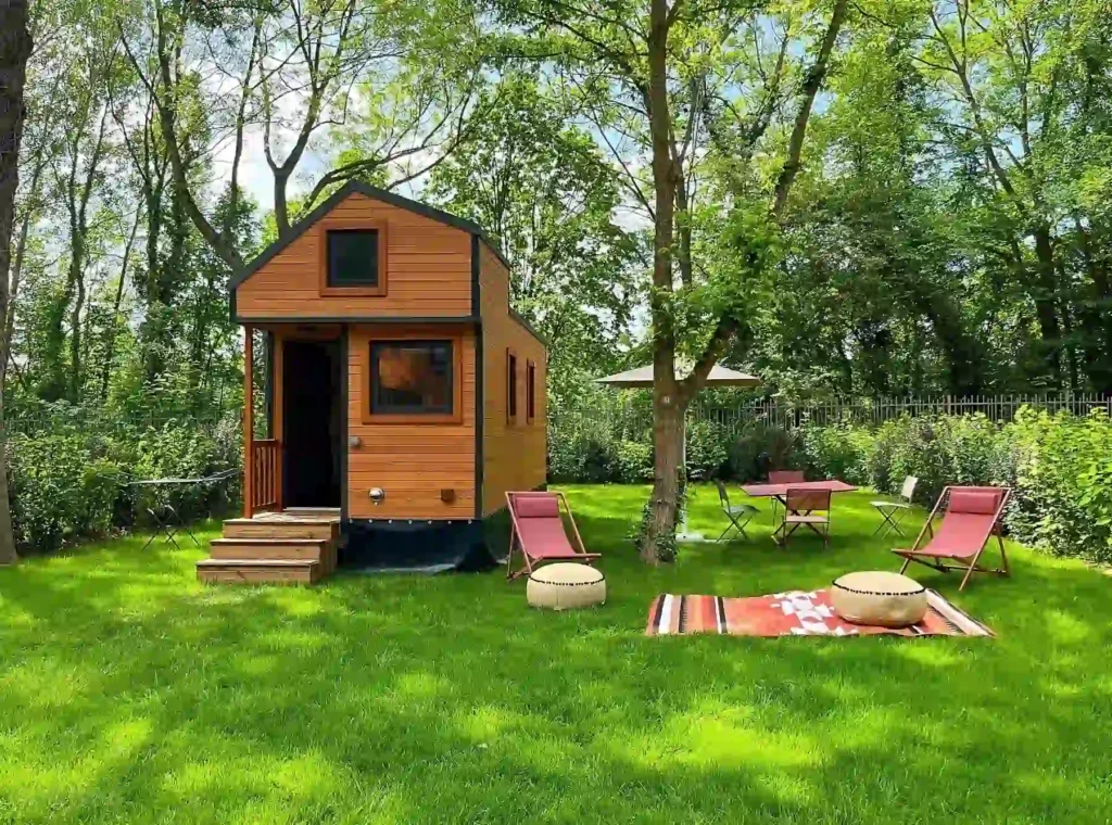 The image depicts a small wooden tiny house set in a lush green backyard surrounded by tall trees. The house has a modern design with natural wood paneling and black trim, a small porch with stairs leading up to the entrance, and a loft-style window at the top. The yard features outdoor seating with red lounge chairs, a small table with a parasol, and a colorful rug laid out on the grass with two cushions for a cozy, relaxing atmosphere. The surrounding environment appears peaceful and secluded, with dense foliage and greenery in the background.