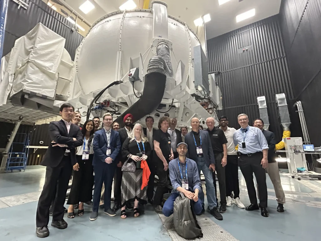 A group of approximately 17 people poses for a photograph in front of a large, industrial, spherical structure, likely related to science or space exploration due to the high-tech equipment in the background. The group consists of a mix of men and women, dressed in both business and casual attire. Most individuals are standing, while one person kneels in the center. They are inside a facility with high ceilings and specialized machinery, and several members are wearing lanyards, possibly indicating participation in a formal event or tour. The lighting is bright and industrial, reflecting the technical setting of the room.