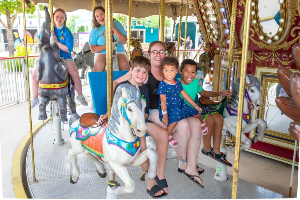 A group of children and an adult are enjoying a carousel ride. The adult is seated in the middle of the carousel with two children on her lap, both smiling brightly. Surrounding them, two older children are riding carousel animals, including an elephant and a horse, while smiling and laughing. The scene is lively and full of joy, with colorful, elaborately decorated carousel animals in the foreground, and a sunny amusement park setting in the background.