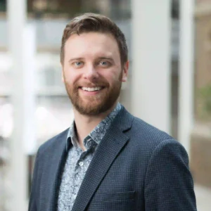The image shows a man with a light complexion, short brown hair, and a beard. He is wearing a patterned shirt and a dark-colored blazer with a subtle checkered pattern. The background appears to be indoors, with soft lighting and a blurred view of what seems like windows or large glass panes. Alt text: "A smiling man with short brown hair and a beard, wearing a patterned shirt and a dark checkered blazer, standing in an indoor setting with blurred glass windows in the background.