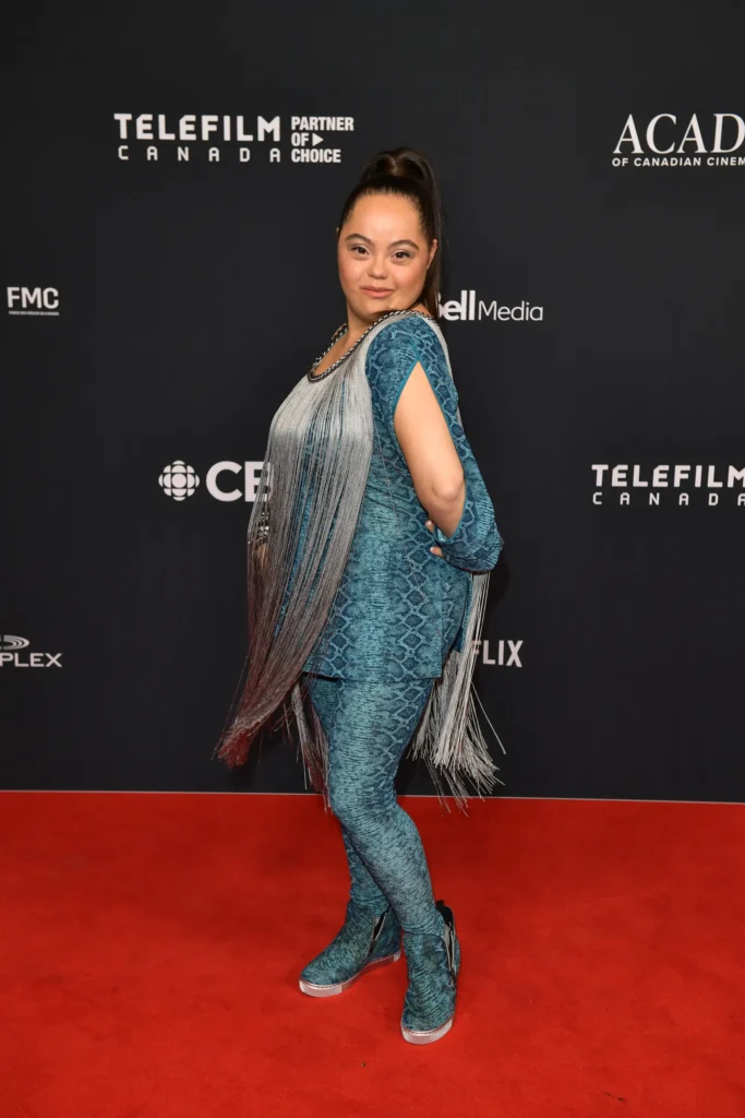 A woman poses confidently on a red carpet in front of a backdrop featuring logos from Telefilm Canada, the Academy of Canadian Cinema, and other sponsors. She is wearing a vibrant blue outfit with a snake-skin pattern, consisting of matching leggings and a tunic. The tunic is adorned with long, silver fringe details that add movement and texture. She is also wearing matching blue ankle boots. Her hair is styled in a high ponytail, and she smiles while striking a bold, stylish pose with one hand on her hip.