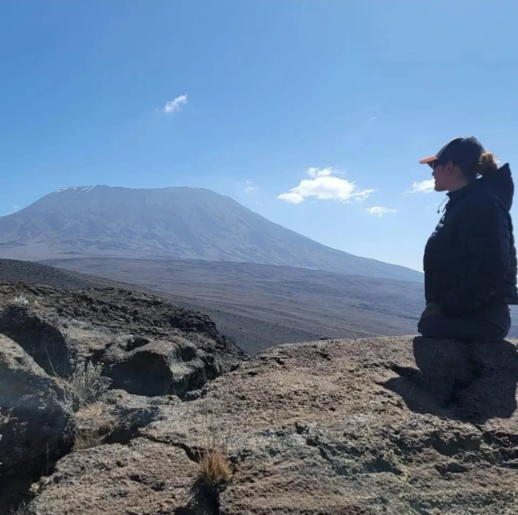 A woman with no legs is seated on a rocky outcrop, gazing towards a distant, towering mountain under a clear blue sky. She is dressed warmly in a dark jacket and a cap, suggesting a cool climate. The landscape around her is barren and rugged, with the mountain dominating the background, possibly a volcanic peak. The atmosphere is serene, emphasizing the vastness of nature and the solitude of the moment