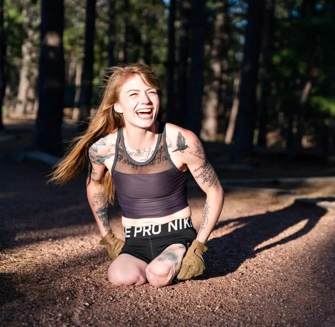 A smiling woman is sitting on the ground in an outdoor forest setting with sunlight filtering through the trees. She has long brown hair and is wearing a black sleeveless crop top and Nike Pro shorts. She is also wearing gloves. She has multiple tattoos on her arms, shoulders, and upper chest. Her legs are amputated above the knees, and she is sitting directly on the ground, beaming with joy, giving off an energetic and positive vibe. The background consists of tall trees and a natural landscape.