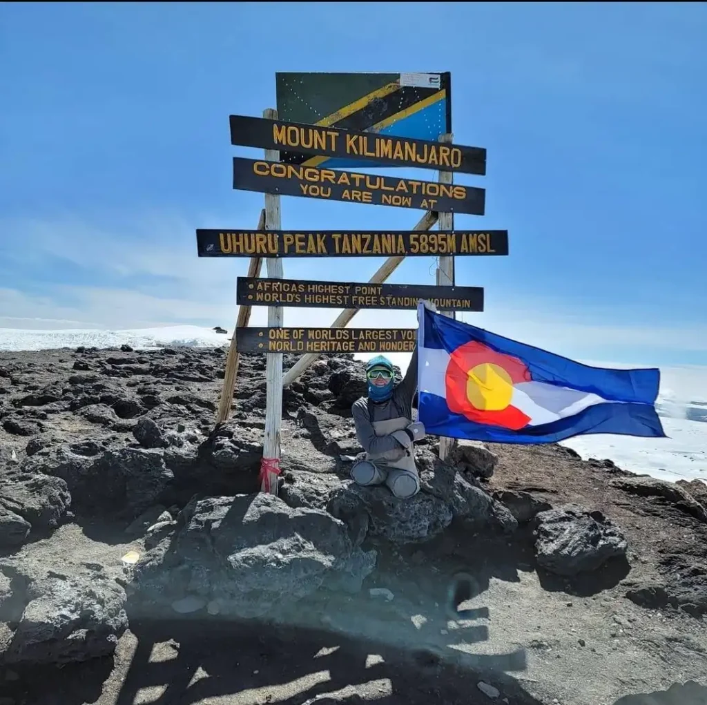 A person standing at the summit of Mount Kilimanjaro at Uhuru Peak, Tanzania (5895m above sea level). They are holding a Colorado state flag and are dressed in mountain climbing gear, including goggles and a face covering. The peak is marked by a wooden sign with several inscriptions, including “Africa’s highest point” and “World’s highest free-standing mountain.” The background shows rocky terrain and snow with a clear blue sky.
