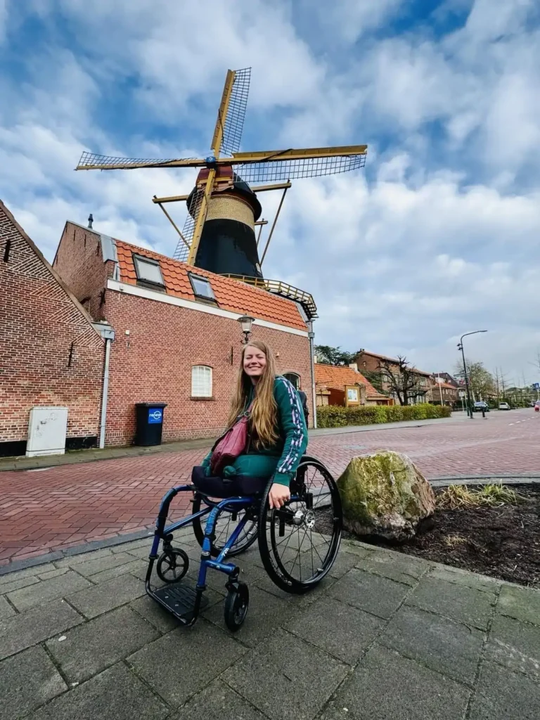 A woman in a wheelchair is smiling and posing in front of a traditional Dutch windmill. She has long hair and is wearing a green tracksuit with a pink bag across her chest. The windmill is large, with classic blades, and the brick building next to it has a tiled roof. The scene takes place on a quiet street, with overcast skies and some greenery around, giving a peaceful, quaint atmosphere. The image highlights accessibility, travel, and enjoyment of cultural landmarks.