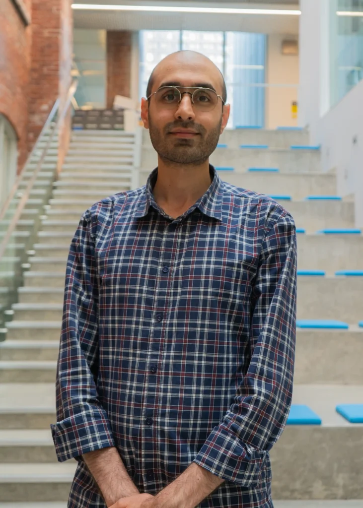 The image shows a man standing indoors, in front of a modern staircase. He is wearing glasses and a checkered shirt with a blue and red pattern. The background features a large staircase with blue accents on the steps, as well as some exposed brick walls and glass panels. The lighting is bright, suggesting the space may be well-lit by natural or artificial light. The man is looking directly at the camera with a neutral expression.