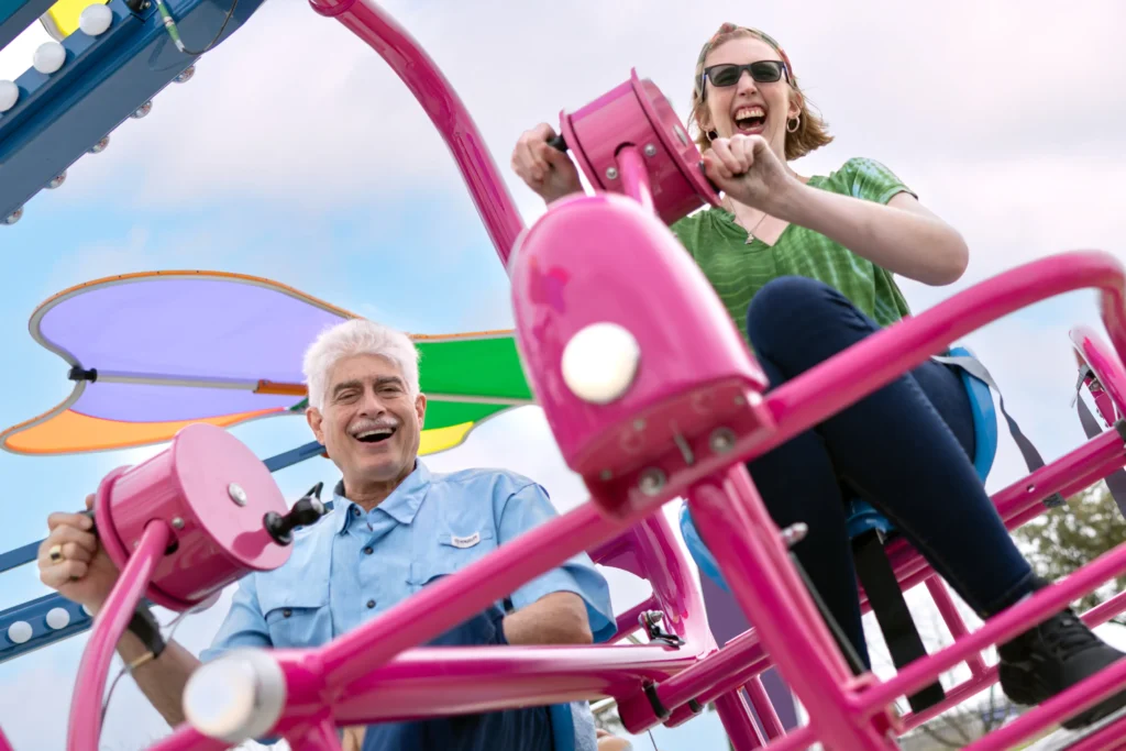 A vibrant image shows two people joyfully riding a colorful amusement bike, featuring bright pink handlebars and frame. The man on the left, with white hair and a big smile, is wearing a light blue shirt, while the woman on the right, also laughing and wearing sunglasses, is dressed in a green shirt with dark pants. Both are energetically turning the cranks of the ride. The background has playful shades of purple, green, and orange from the canopy above, adding to the cheerful and lively atmosphere of the scene.
