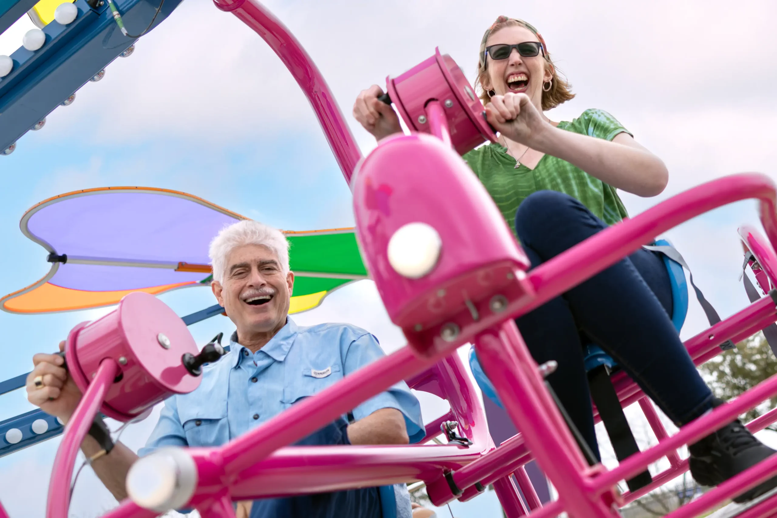 A vibrant image shows two people joyfully riding a colorful amusement bike, featuring bright pink handlebars and frame. The man on the left, with white hair and a big smile, is wearing a light blue shirt, while the woman on the right, also laughing and wearing sunglasses, is dressed in a green shirt with dark pants. Both are energetically turning the cranks of the ride. The background has playful shades of purple, green, and orange from the canopy above, adding to the cheerful and lively atmosphere of the scene.