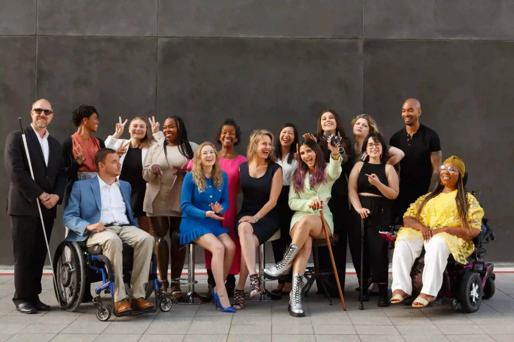 A diverse group of fifteen people pose together, smiling and laughing, showcasing a variety of expressions, poses, and personalities. The group includes individuals with visible disabilities, such as wheelchairs, canes, and prosthetic limbs. The members represent various ages, genders, and ethnicities, reflecting a strong sense of inclusivity and camaraderie. The background is a plain, dark wall, which brings focus to the group in the foreground, and the image captures a joyful, empowered atmosphere.