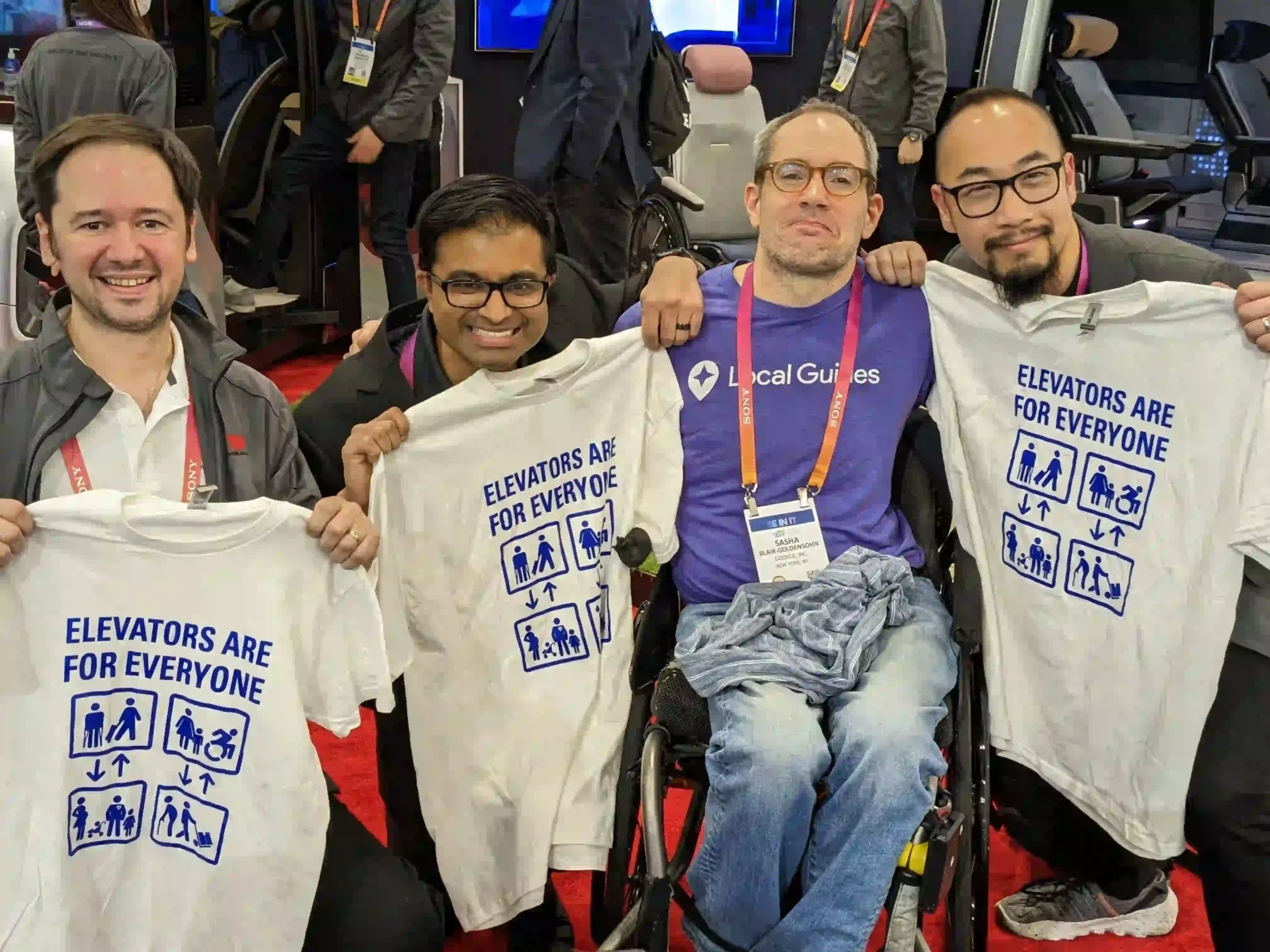 Four people are posing in the image. One of them, seated in a wheelchair, wears a purple "Local Guides" shirt, while the other three stand around them, smiling. Two of them are holding up white t-shirts that say "ELEVATORS ARE FOR EVERYONE" in blue, accompanied by icons representing accessibility for people of all abilities. The group appears to be in a public or conference setting, with red carpeting and visible lanyards on some of the individuals, suggesting they are participating in an event.