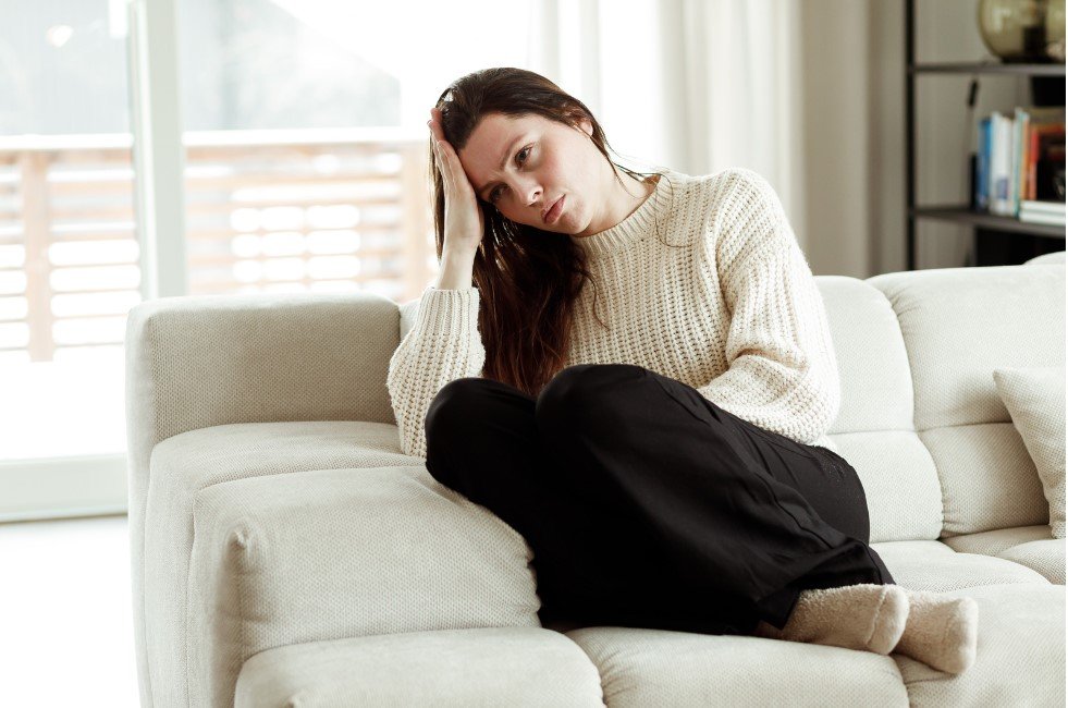 The image shows a woman sitting on a beige couch, leaning slightly to one side with her hand resting on the side of her head. She appears to be deep in thought or feeling tired. She is dressed casually in a cream-colored knitted sweater and black pants, with her legs crossed and wearing cozy socks. The setting looks like a bright and modern living room, with light streaming in from a window or door in the background.
