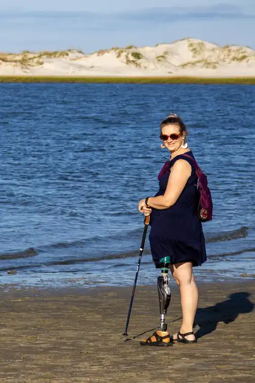 The image shows a woman standing on a beach near the water, smiling and facing the camera. She has a prosthetic leg and is using a walking stick for support. She is wearing sunglasses, a dark sleeveless dress, and sandals. The background features calm water and sand dunes under a clear sky, creating a peaceful beach scene.