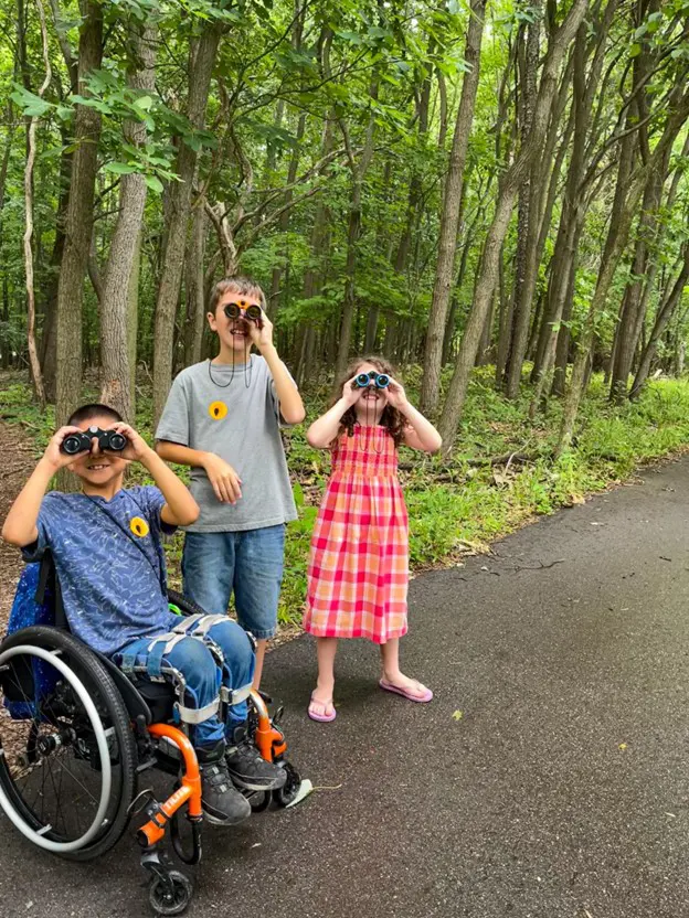 The image shows three children engaging in birdwatching or nature observation using binoculars. They are standing on a paved path in a wooded area. One child is seated in a wheelchair, while the other two stand beside them. All three appear to be looking up into the trees with curiosity. The setting is a forest or park with tall trees and greenery surrounding the path. The children are dressed casually, and their expressions suggest they are focused and enjoying the activity.