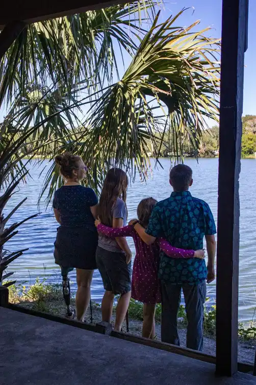 A family stands by a lakeside, viewed from behind under the shade of a large palm tree. There are four people in the image: an adult on the left, who has a prosthetic leg, and three children beside them. The two adults, one child in the center, and one on the right are all facing the lake, enjoying the view. The child on the right has their arms around the others, creating a sense of togetherness. The scene is peaceful, with a serene body of water in the background and bright daylight filtering through the palm leaves.
