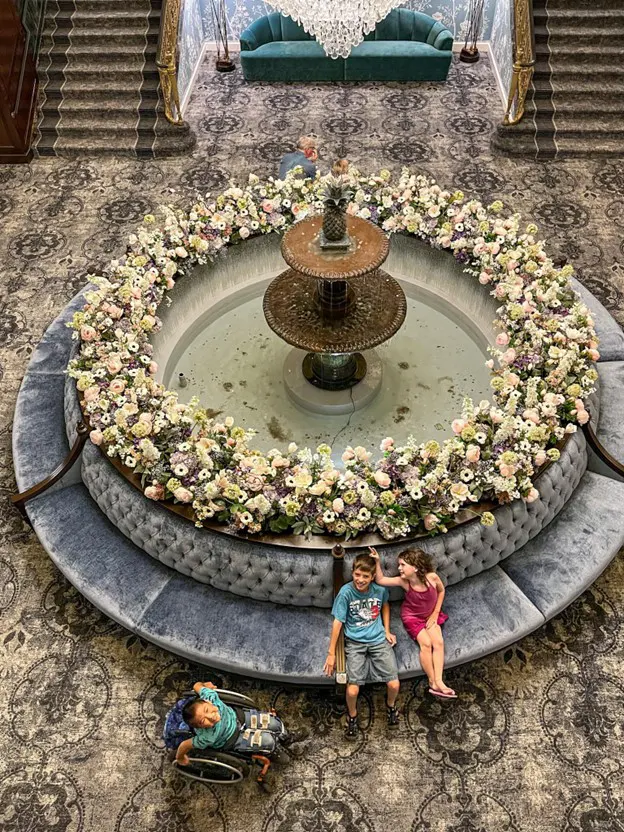 The image depicts an ornate interior setting, with a large circular fountain at the center, adorned with an abundance of pastel-colored flowers along its edge. Surrounding the fountain is a plush, circular, tufted sofa in a greyish-blue hue. Two children, a boy and a girl, are seated on the sofa, smiling and interacting with each other. Another child in a wheelchair sits nearby, gazing toward the center. In the background, there is a wide staircase covered in patterned carpet, leading up to a second level, where a greenish-blue couch is visible beneath a grand chandelier. The overall atmosphere feels elegant and serene, with intricate details in both the decor and furnishings.